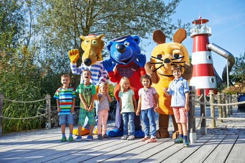 Ravensburger Spieleland (© Ravensburger AG). 7 Kinder stehen zusammen mit den Kinderfilmfiguren "Hein Blöd", "Käptn Blaubär" und der "Maus" auf einem Holzsteg vor einem Rutschturm im Ravensburger Spieleland