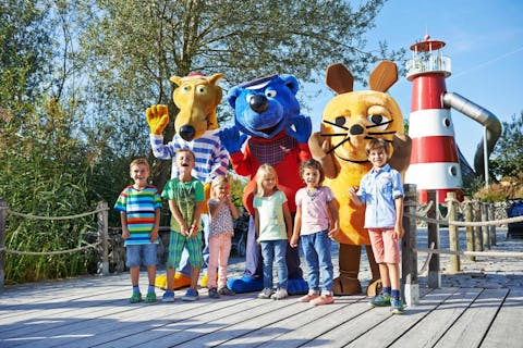 Ravensburger Spieleland (© Ravensburger AG). 7 Kinder stehen zusammen mit den Kinderfilmfiguren "Hein Blöd", "Käptn Blaubär" und der "Maus" auf einem Holzsteg vor einem Rutschturm im Ravensburger Spieleland