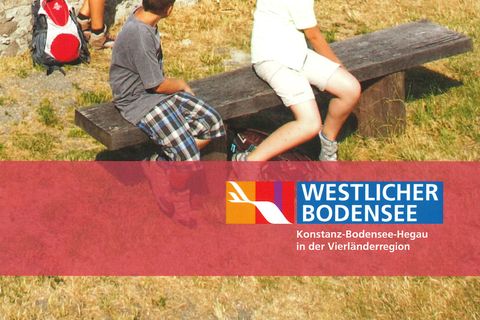 Titelseite Wanderkarte "Hegau-Panoramaweg". Auf dem Bild ist eine Frau (an der Mauer stehend) mit 2 Kindern auf einer Bank sitzend zu sehen, die vom Hohentwiel über die Stadt Singen Singen und den Hegau schauen.