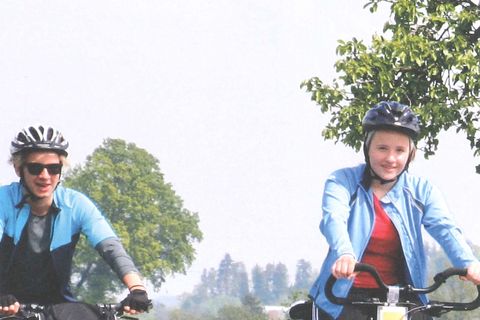Titelblatt Flyer MeinRadweg. Ein junges Pärchen fährt mit dem Fahrrad durch eine grüne Wiese mit gelben Sumpfdotterblume. Oben ist das Logo ist der Schriftzug "Meinradweg" mit dem Logo zu sehen. Das Logo ist ein Mönch auf einem Fahrrad. 