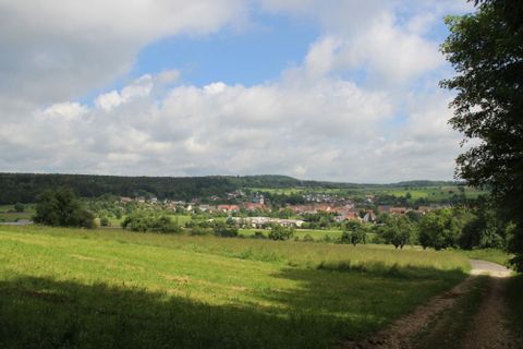 Wanderweg Südwind - Blick aus dem Wald heraus in die freie Landschaft mit grünen Wiesen und Bäumen und der Ortschaft Heudorf im Hintergrund. Der Himmel ist sonnig, aber auch wolkig..