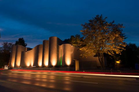 Das geschwungenge, erdfarbene und beleuchtete Gebäude des MAC Museum Art & Cars, das von von seiner Architektur den Hohentwiel und die Burgruinie darstellen soll,  von der Außanansicht von Richtung Schaffhauser Straße  (© MAC Museum Art & Cars Singen)