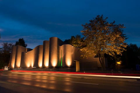 Das geschwungenge, erdfarbene und beleuchtete Gebäude des MAC Museum Art & Cars, das von von seiner Architektur den Hohentwiel und die Burgruinie darstellen soll, von der Außanansicht von Richtung Schaffhauser Straße (© MAC Museum Art & Cars Singen)