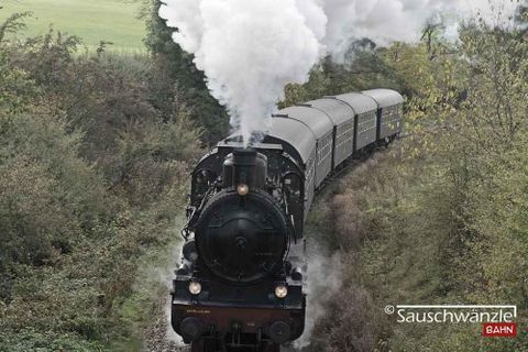 Die nostalgische Sauschwänzlebahn fährt stilgerecht mit einer historischen Dampf- und Diesellok mit fünf, schwarzen Waggons auf Schienen durch die Landschaft. Aus dem Schornstein der Lok steigt viel weißer Rauch auf. (@Sauschwänzlebahn)