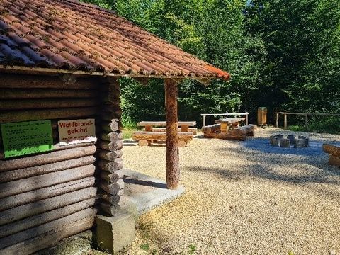 Hier ist der Grillplatz an der Reschberghütte zu sehen. Neben der Grillstelle sind Sitzmöglichkeiten und ide angrenzende Reschberghütte, eine Holzhütte, zu sehen.